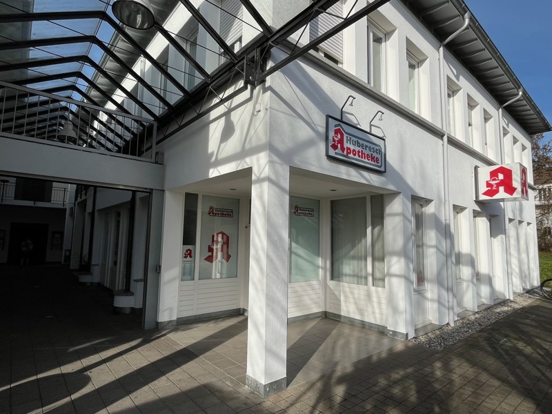 Eingangsbereich der Huberesch Apotheke in Ravensburg mit Glasüberdachung und Apothekenlogo. Helle Fassade mit Beschilderung „Huberesch Apotheke“ über der Eingangstür.